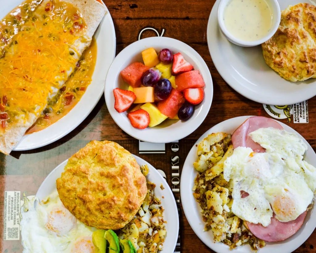 A table at Dot's Diner seen from above, with a smothered burrito under green chili and cheddar, a bowl of cut fruit, a biscuit with a cup of gravy, hash browns under eggs and avocado, and ham and eggs with potatoes.